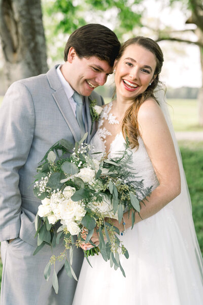 Groom laughs with his bride holding a lush bouquet of white flowers. Joyful wedding captured by Jackson Ms Wedding Photographer.