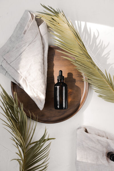 Dropper bottle with natural serum on wooden tray surrounded by palm leaves. 