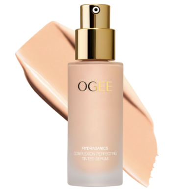 A frosted glass bottle of OGEE Hydraganic Complexion-Perfecting Tinted Serum with a gold pump, featured with a smooth swatch of peach-toned product behind it. Combines skincare and coverage for a radiant finish.