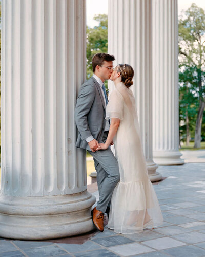 A couple dressed in wedding attire holds hands and kisses between large white columns outdoors, surrounded by trees—captured beautifully by an NJ wedding photographer.