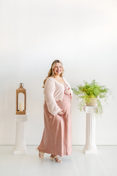 Pregnant woman standing between two white pillars, gently holding her belly and looking at the camera; one pillar holds a lantern, the other a large green plant