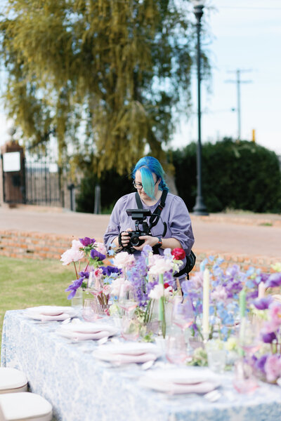 Woman behind a decorated table at a wedding with blue haor looking down at her camera filmming closeups on her camera of the flowers