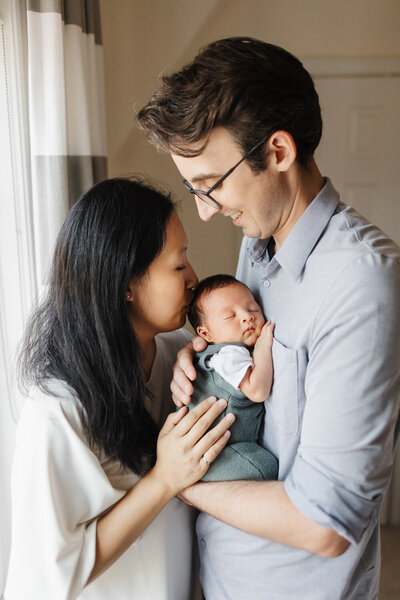 mom giving newborn son a kiss on the head while dad holds him close