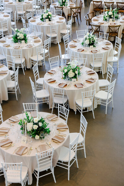 Elegant wedding reception setup featuring round tables draped in ivory linens, white Chiavari chairs, and lush white-and-green floral centerpieces with roses, hydrangeas, and greenery, styled with neutral napkins, gold flatware, and candle accents—modern timeless wedding décor inspiration.