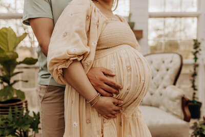 A mother holds her bump with her partner behind her holding her bump from behind. They are posing for San Antonio maternity photography session.