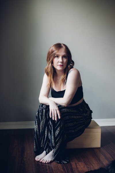 A woman with long red hair sits barefoot on a wooden block, wearing a black top and patterned pants, looking at the camera—the owner of an agency offering branding and web design for female entrepreneurs.