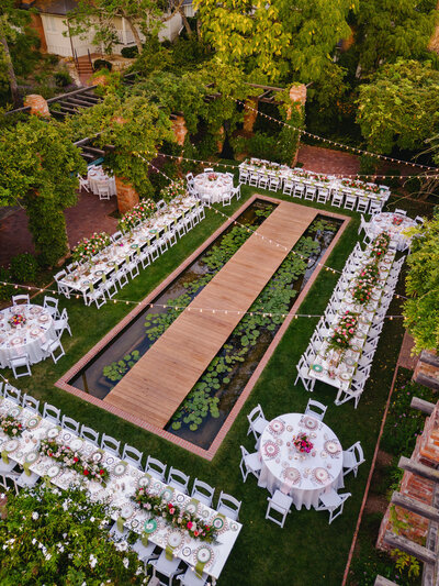 Elegant BP Collective El Encanto Garden Party with round tables set around a lily pond, white chairs, string lights, and lush greenery.