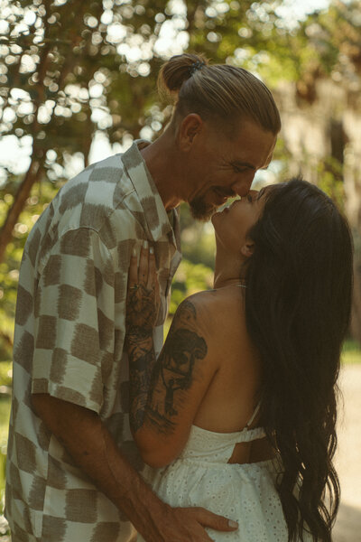 Expecting couple sharing an intimate embrace under a large oak tree during golden hour at a garden in Florida; woman in a white eyelet dress with long dark hair and man in checkered shirt lovingly holding her belly.