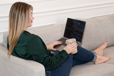 Woman sitting on a couch with a laptop open on her lap and coffee in hand. She is wearing jeans and a long sleeve green shirt.