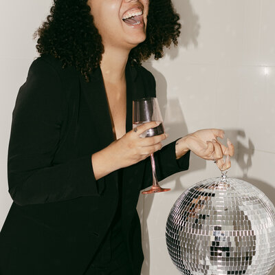 Catryce holding a wine glass and disco ball in a pantsuit