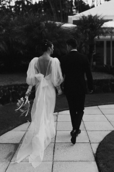 A film portrait of a bride and groom walking hand-in-hand on their wedding day at Puketutu Island Estate. Captured by Eilish Burt Photography