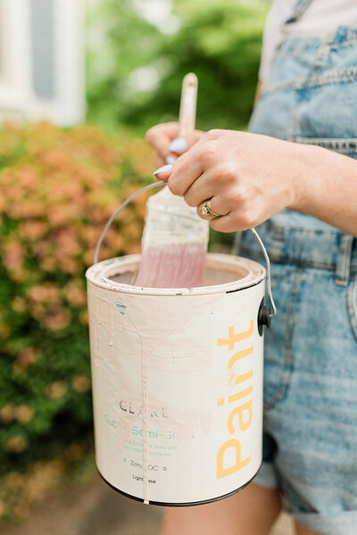 A close up photo of a woman wearing overalls, holding a can of house paint and a paint brush in preparation for a diy project.
