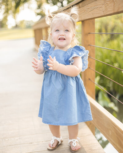 Toddler girl with blonde pigtails, blue and white dress, on a bridge at a wildlife sanctuary, looking away from the camera with a smile and starting to clap