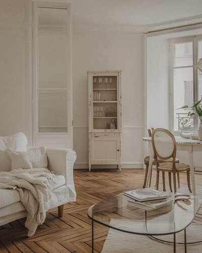 patterned wooden floor with white room white furniture, glass coffee table with books and tan caned dining chairs