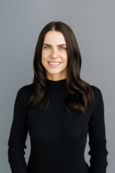 A woman with dark hair, wearing a black ribbed sweater, smiles against a neutral grey background. A clean and timeless professional headshot.