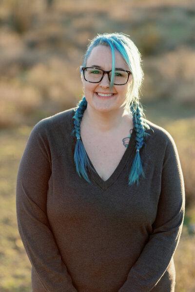 Woman smiling in a grey sweater with blue hair in french braids wearing glasses.