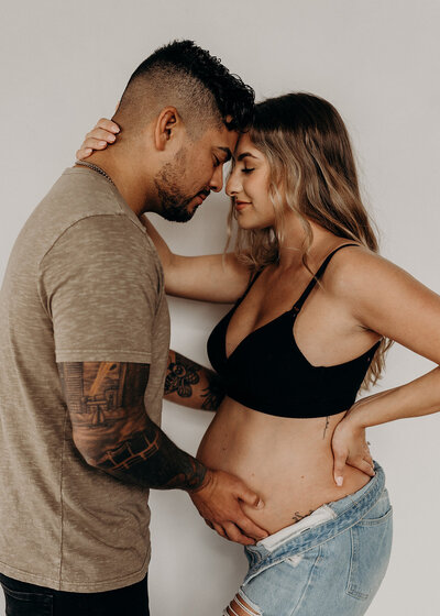 A multi racial couple stands facing each other with their foreheads together while the tattooed husband holds his wife's bear pregnant belly while she wraps her arms around his neck in and bright and airy photography studio in Denver, Colorado.
