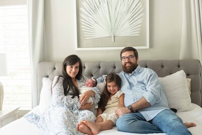 Family of four sits on a bed smiling, with mom holding a newborn and big sister leaning in close.
