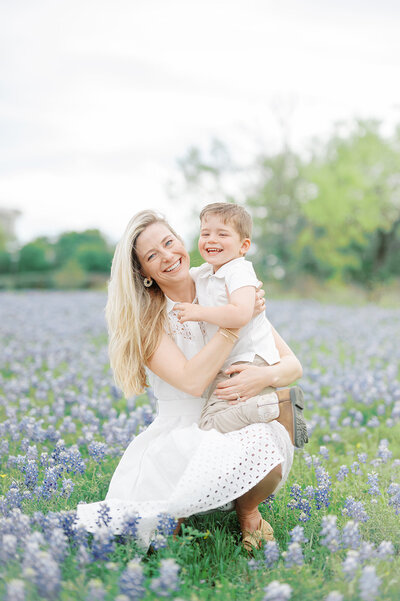 A joyful mother embracing her young son while kneeling in a picturesque field of Texas bluebonnets, showcasing natural smiles and loving connection. Perfect for spring family photography in Dallas.
