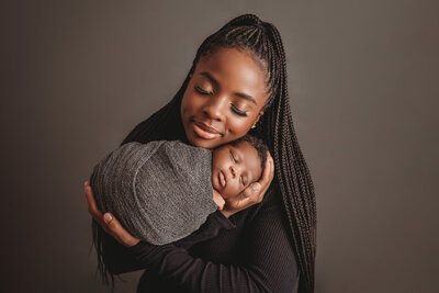 Mom and baby posing for newborn portrait on gray backdrop with mom snuggling baby boy close to her face, both have eyes closed