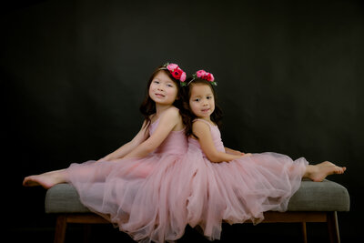 Two sisters in pink tutus sit back to back smiling at the camera