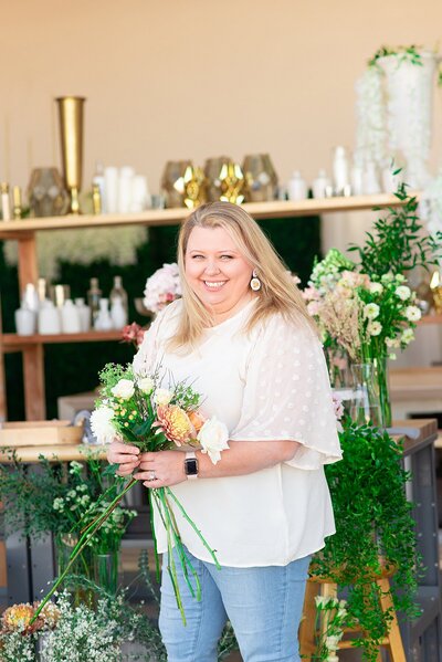 Florist wearing cute cream top and holding bouquet while smiling at camera in her flower shop