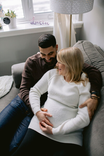 A loving couple sits on a cozy couch, embracing and holding the mother’s baby bump as they share a quiet, intimate moment. The warm, homey atmosphere reflects their journey to parenthood and the deep connection they share. Captured in a natural, heartfelt style by KCP Families