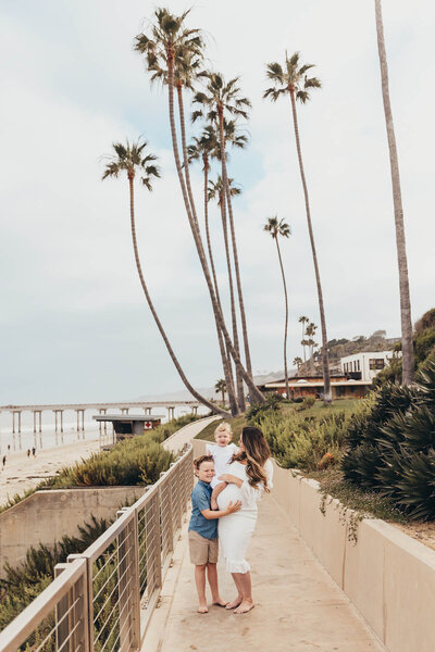 A mother holds her young daughter while her son hugs her baby bump with palm trees in the back