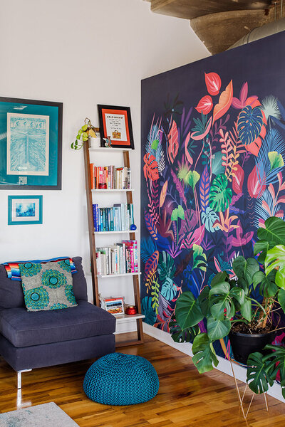 A cozy reading corner with a dark blue chaise lounge adorned with a colorful patterned pillow and a striped throw. A teal knitted pouf sits in front. A leaning wooden ladder shelf filled with books and small decorative items stands against a white wall with two framed pictures above the chaise. The adjacent wall features a vibrant, colorful tropical leaf mural. A large potted monstera plant sits on the wooden floor in the corner.