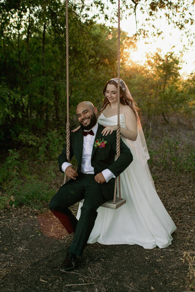 A bride in a white dress and veil stands behind a groom seated on a swing in a forest. The groom wears a dark suit with a bow tie. They smile warmly, surrounded by greenery, with sunlight filtering through the trees.