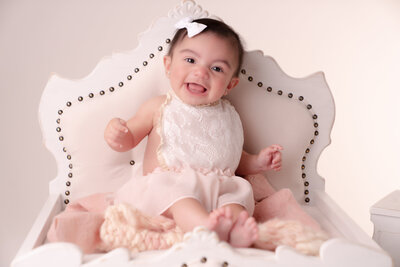 Smiling baby in a white dress and headband sits on an ornate, padded chair. The setting is soft and warm, conveying a joyful and gentle atmosphere.