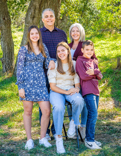Dressed in palette of blues and purples, a family of five stands  and sits together close for a portrait with a backdrop of green grass and sun-lit trees. Photo by SAVI Photography -  Photographer in Riverside