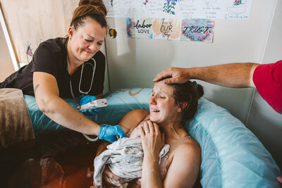 mother holds her baby and cries just after birth with midwife smiling and father's hand on her head