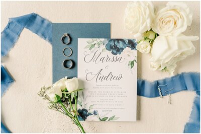 white and blue wedding invitation flat lay with white roses around the invitation and laying on top of blue ribbon