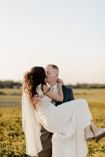 Groom carries his bride and kisses while taking couple portraits during their outdoor wedding ceremony