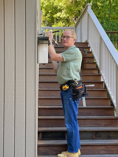 A contractor installing an exterior light fixture on a staircase landing, wearing work gloves and tool belt during a home improvement project.