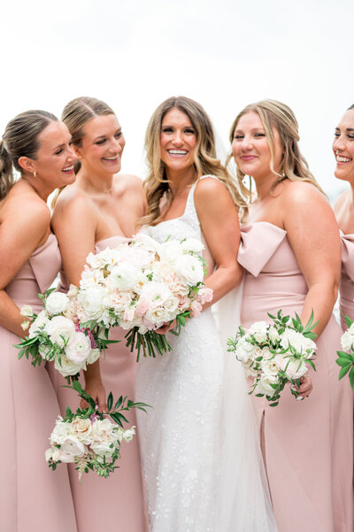 A bride surrounded by her bridesmaids in matching blush pink dresses, all holding soft white and pink floral bouquets, captured at a coastal wedding at The Bohlin in Newport, RI.