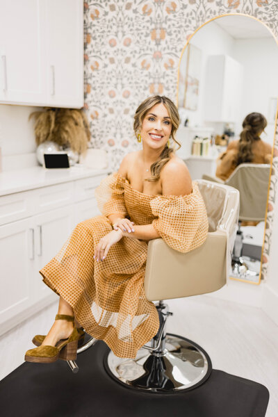 woman sitting in a hair stylist chair wearing a loose dress while smiling