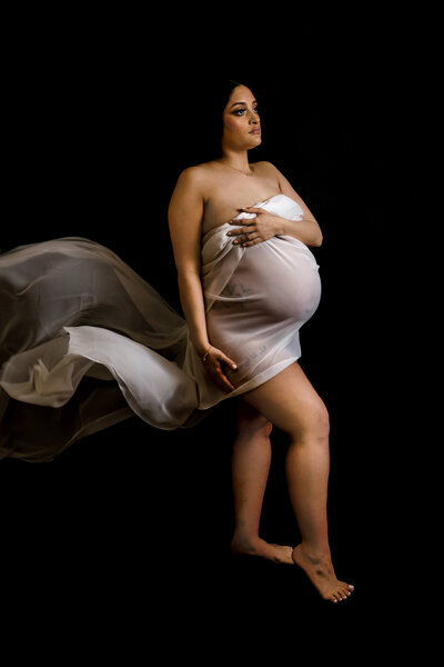 Expecting mother in flowing champagne fabric wrap poses dramatically against black backdrop during artistic studio maternity session with Connecticut maternity photographer