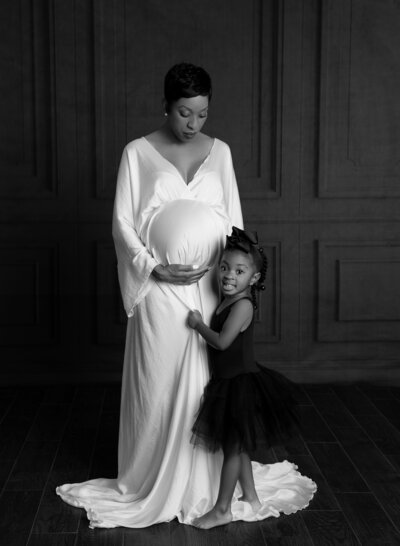 Pregnant mom wearing white sating gown with toddler wearing black tutu dress
