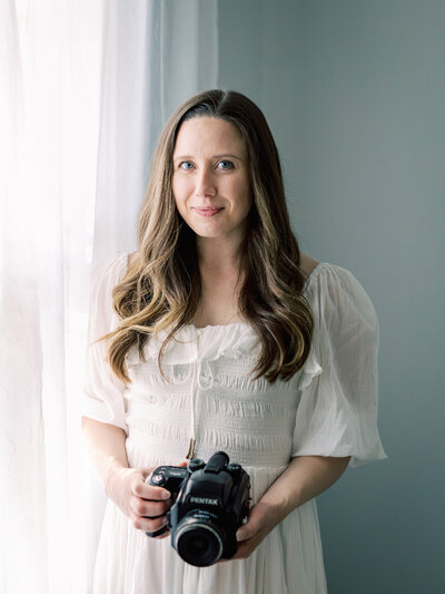 A woman in a white dress standing by a window and holding a camera by Katie Stansfield Photography.