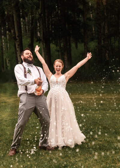 groom popping a bottle of champagne as his bride cheers him on