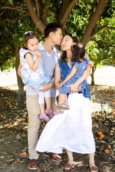 A sweet family of four shares a tender moment under orange trees, captured by Ellobelle Photography in soft, golden light.