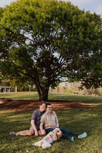 A couple sits with their dog under a tree during their Austin engagement photo session at Mueller Lake Park