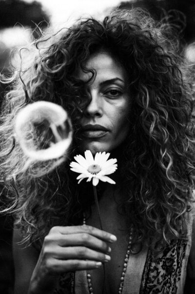 In a captivating black-and-white portrait, the woman with curly hair embodies Sweet Pea Ink Creative's brand identity as she gently holds a daisy while a bubble drifts gracefully in the foreground.
