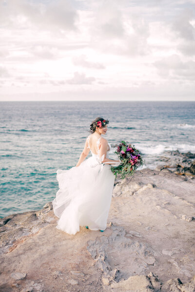 bride on the cliffs of Maui holding a large bouquet with greenery and burgundy flowers. Bride has a head lei on and a billowing dress.