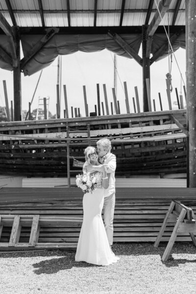 black and white photo of bride and groom in front of boat shed