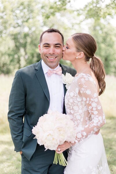 Groom smiling at the camera while bride kissing him on a cheek