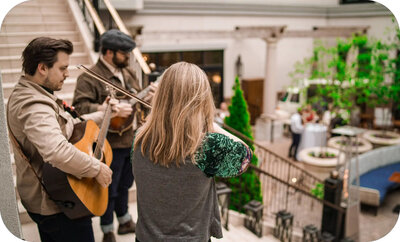Small trio of musicians playing guitar, violin, and another stringed instrument on a staircase overlooking an indoor courtyard.