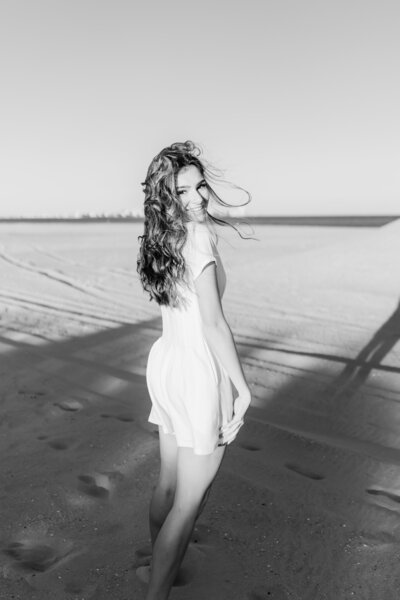 Black and white image of a teen girl dancing on the beach in Ocean City, New Jersey. She is wearing a white romper. and smiling. The sun is casting dark shadows behind her.